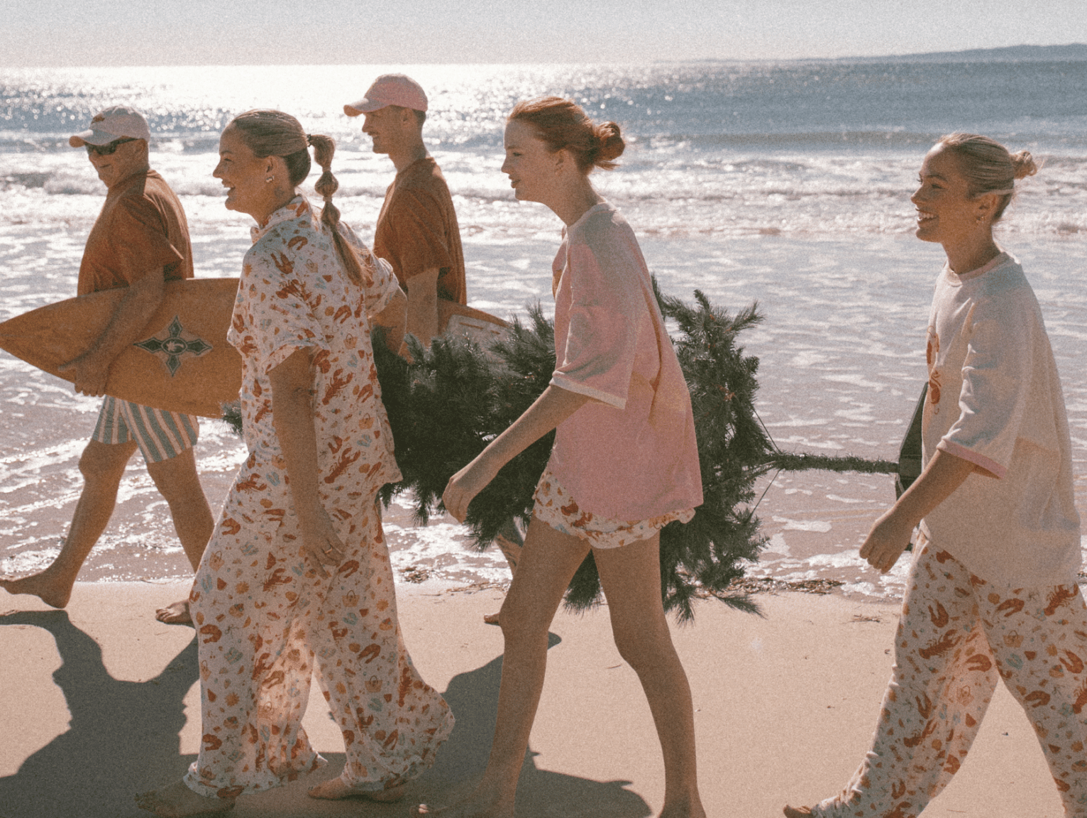 Family walking along the beach wearing christmas pyjamas, holding a christmas tree and surf board.