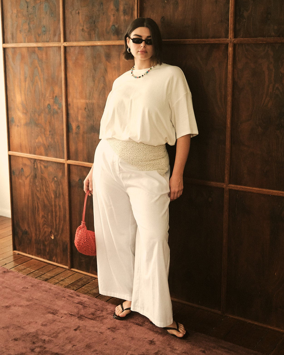 A woman wearing a white short-sleeve top and matching wide-leg pants stands against a dark wood-paneled wall, holding a small red handbag.