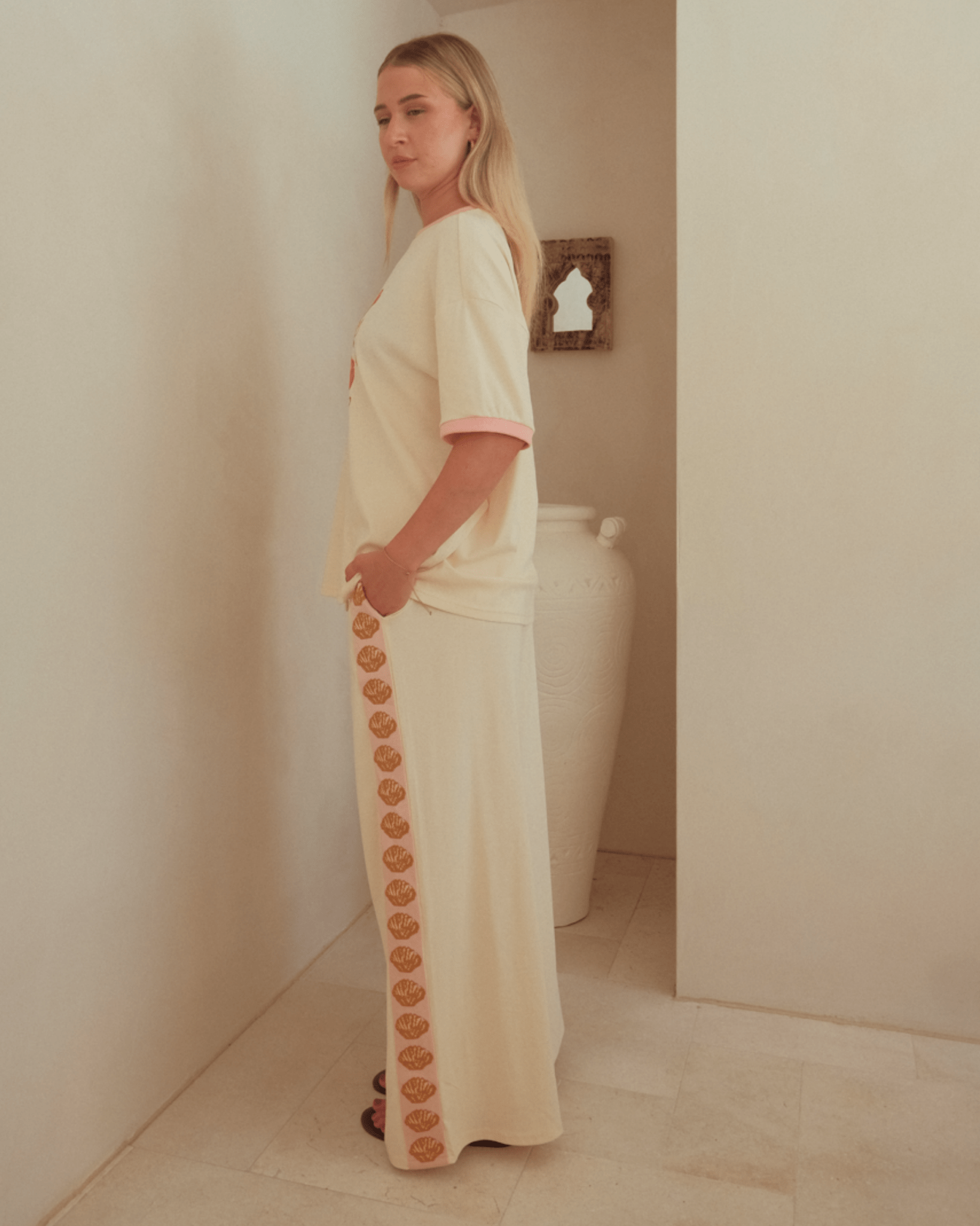 Woman wearing matching beige comfortable lounge set with pink & gold shells down the side-seams of pants.