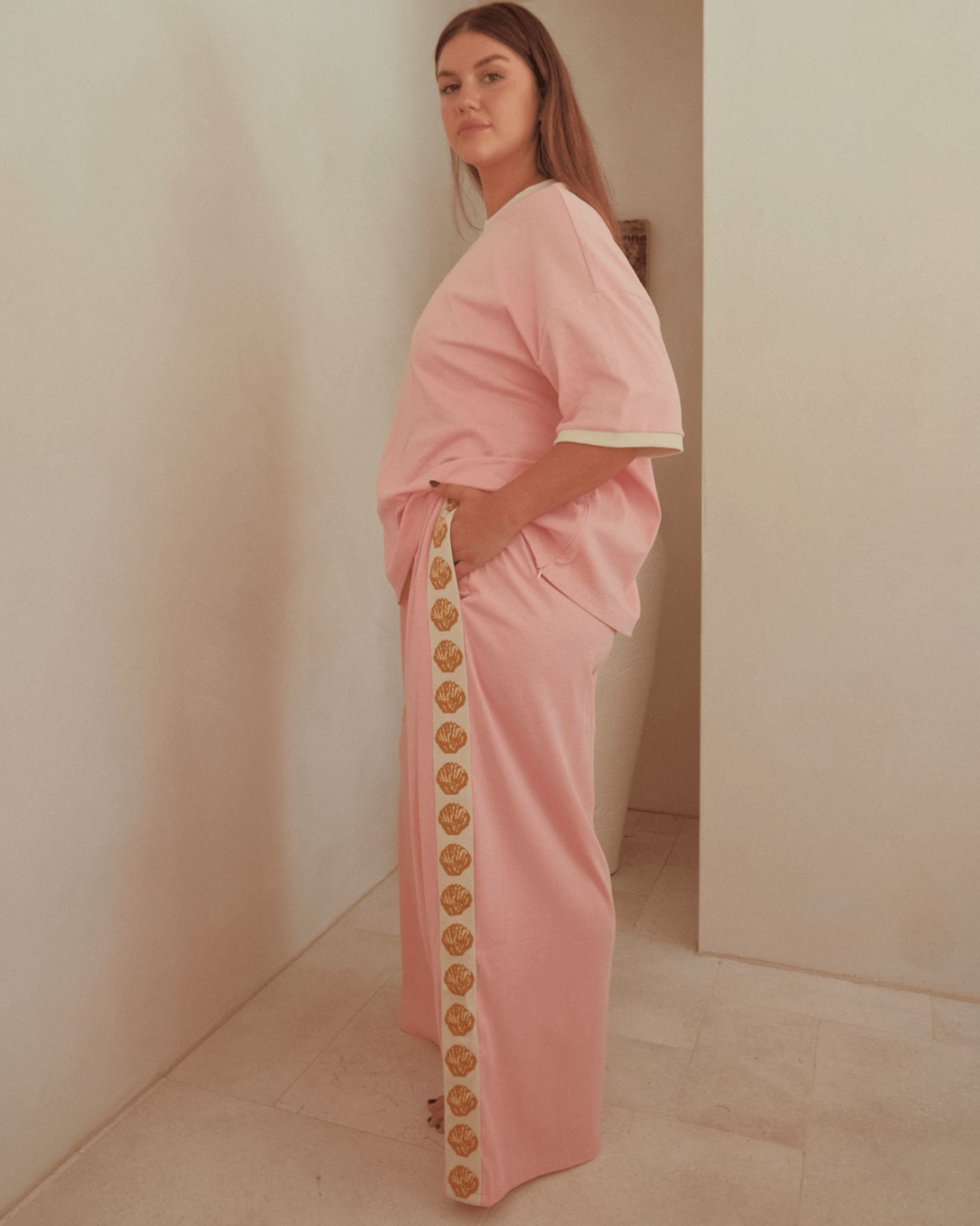 Woman wearing oversized pink shirt and wide-legged pink pants with gold shell detailing down the side. Featuring pockets in the pants.