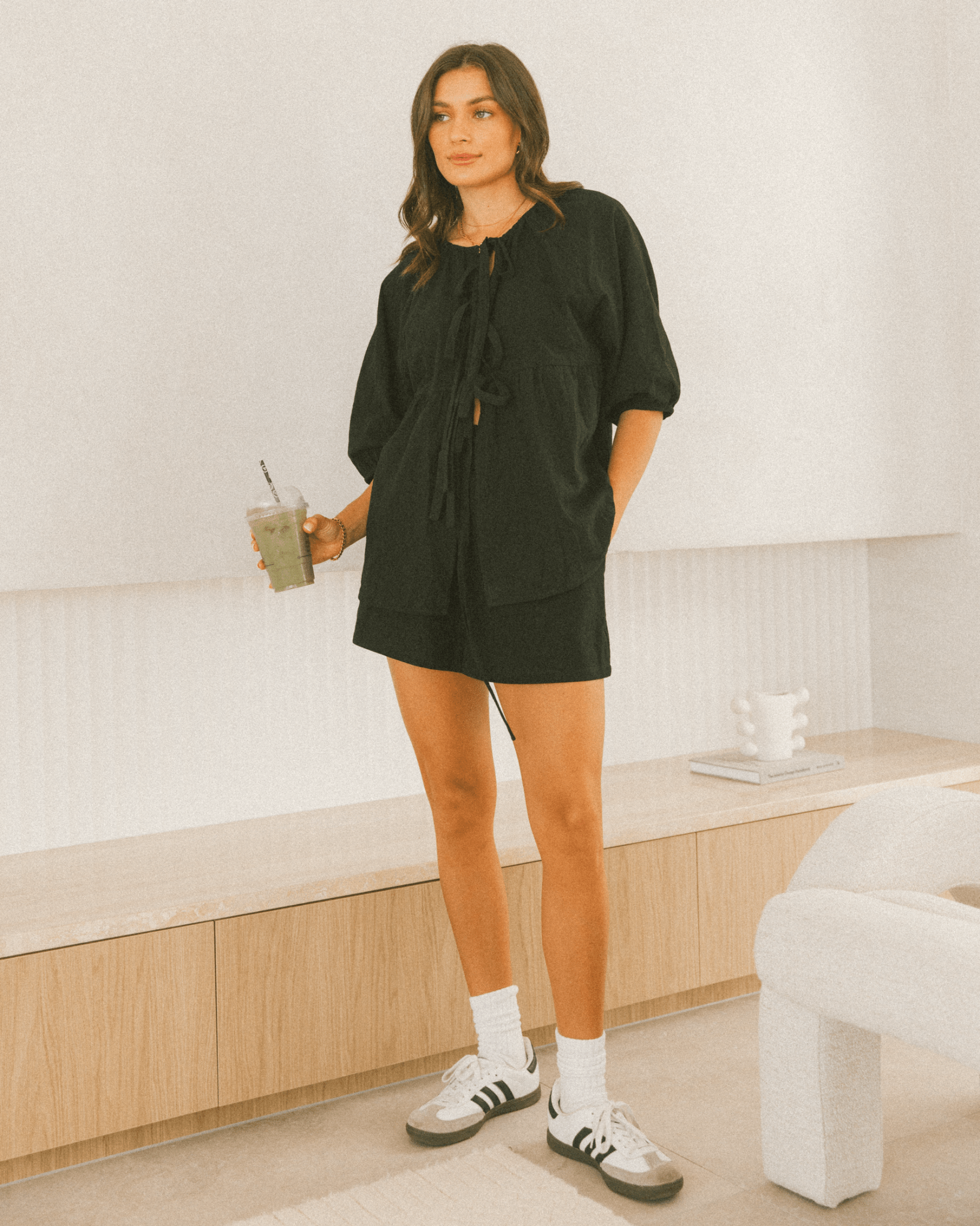 A woman wearing a black oversized top with short sleeves, paired with white sneakers and white socks, standing in a minimalist setting.