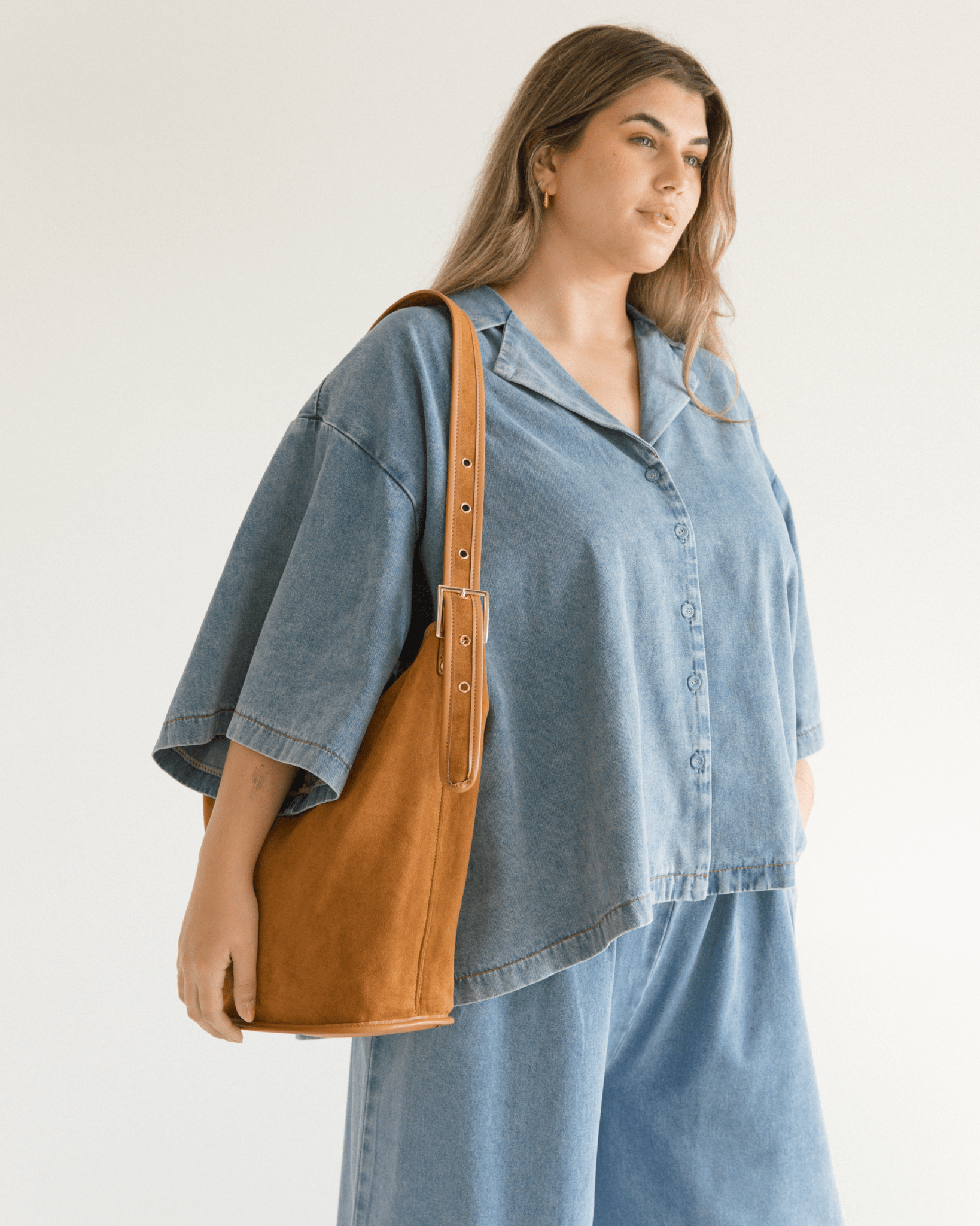 Woman wearing a denim outfit with a brown suede-look bag on a plain background