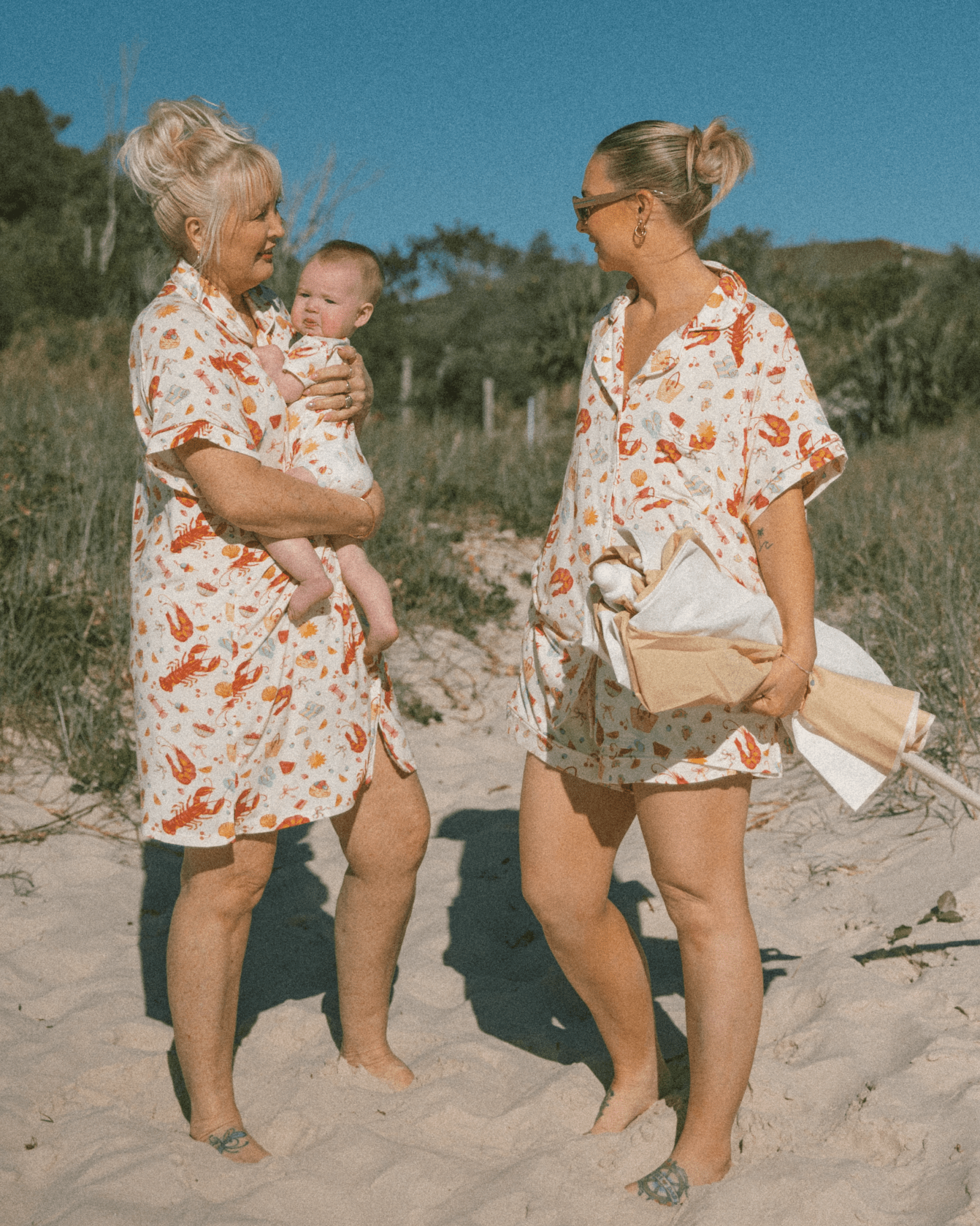 Two women in matching pattern outfits standing on a sandy beach with a baby.