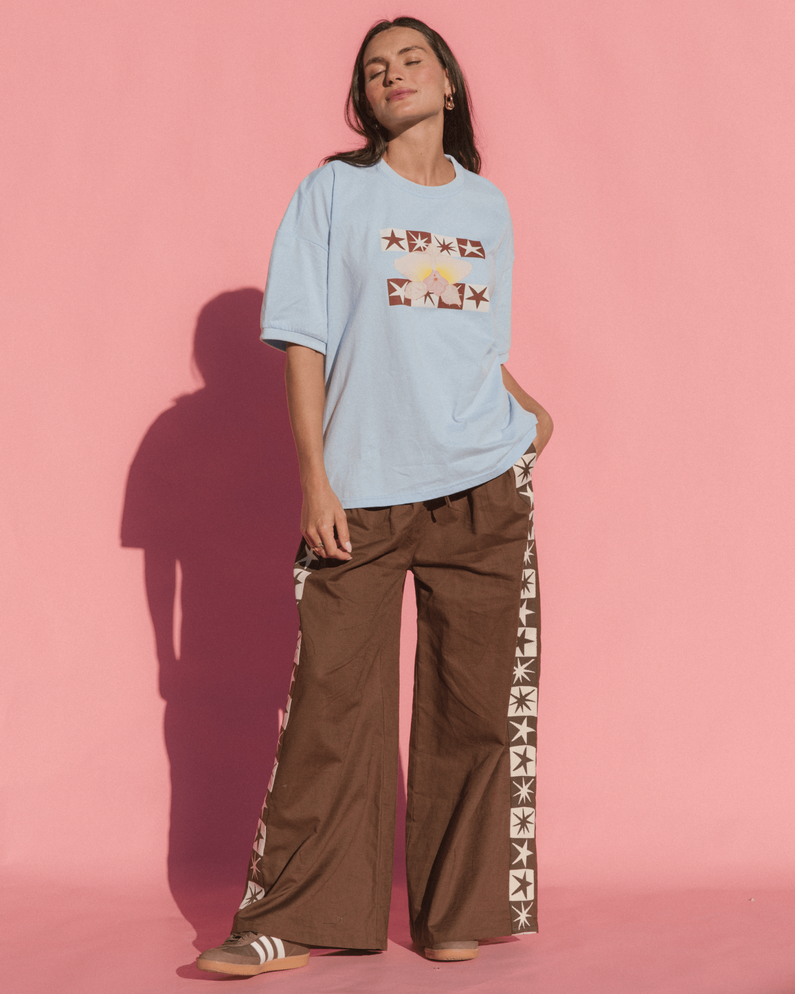 Person wearing a light blue t-shirt with graphic designs and brown pants with star patterns on a pink background
