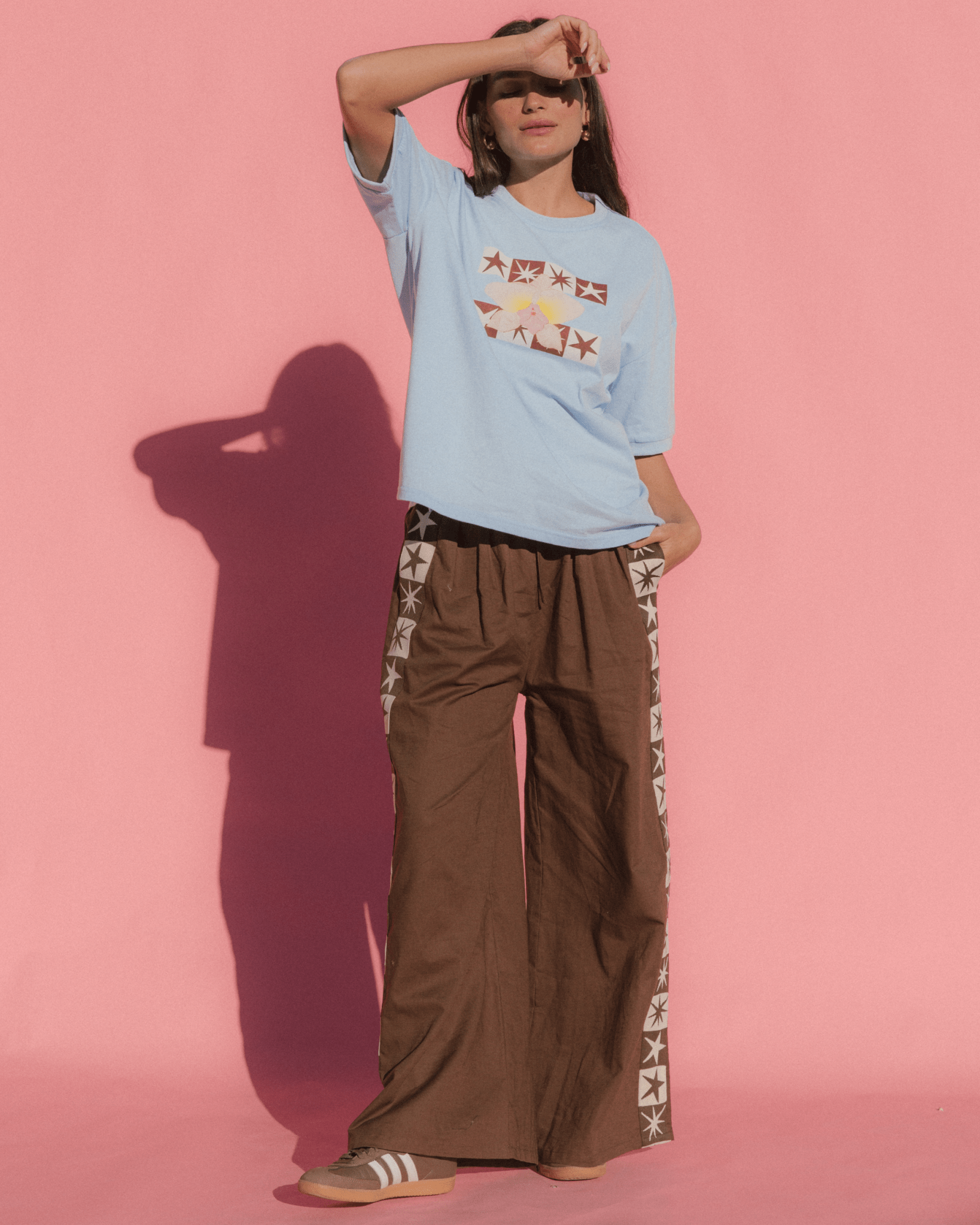 Person wearing a light blue t-shirt with graphic designs and brown wide-leg pants on a pink background