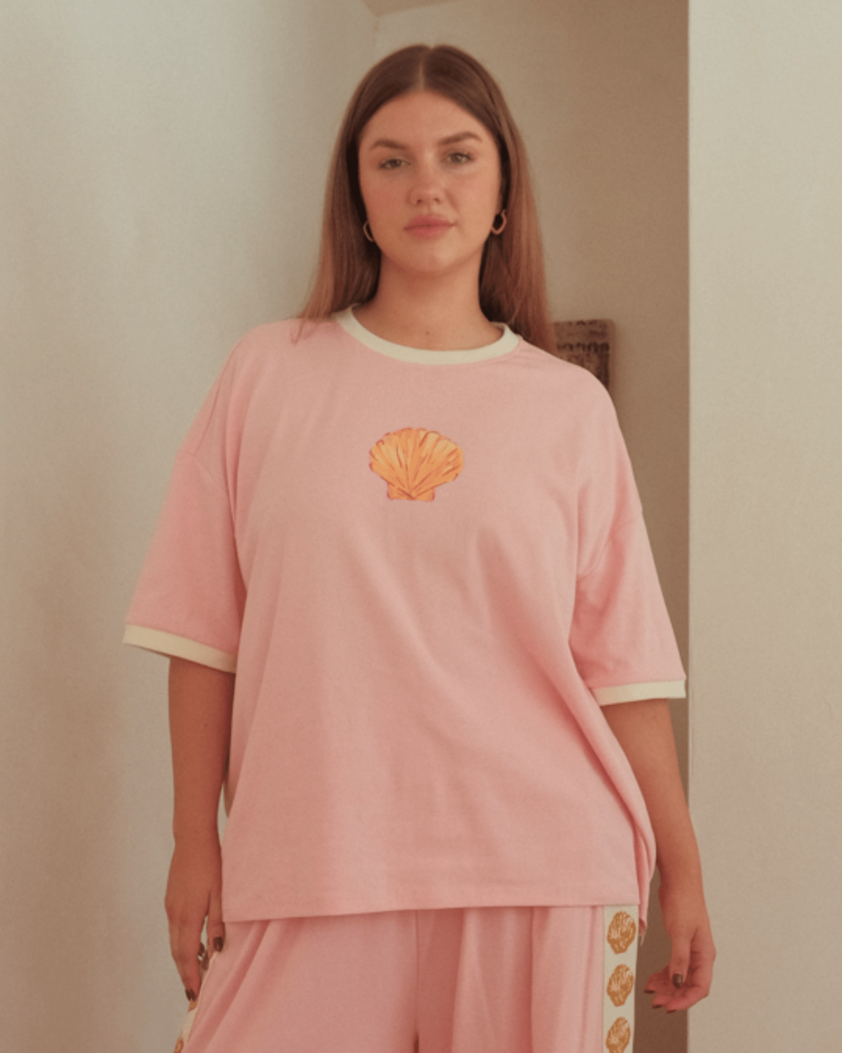 Woman wearing oversized slouch-style pink tshirt with gold shell design on the front.