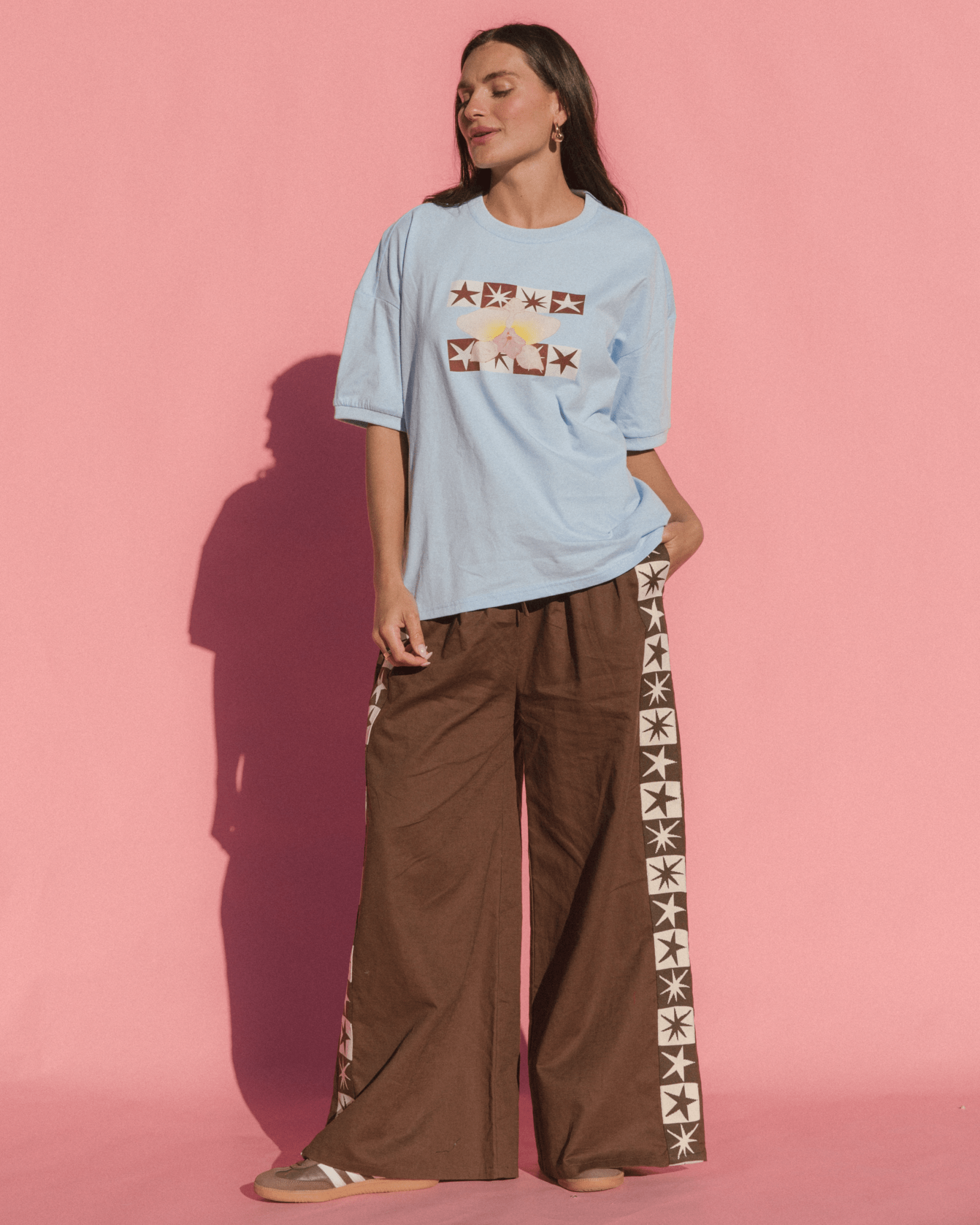 Woman wearing a light blue t-shirt with brown graphic floral & star design and brown pants with star patterns against a pink background