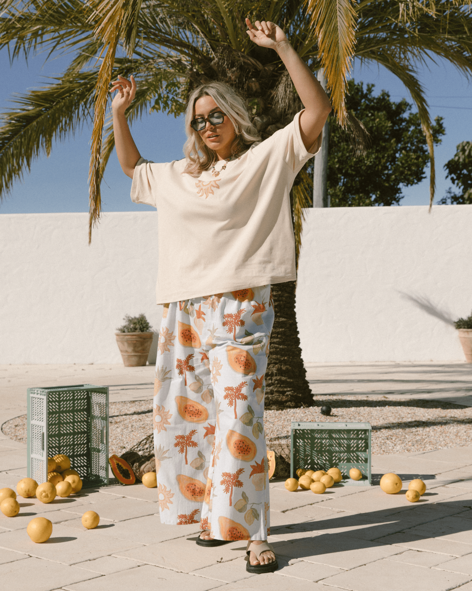 Woman in a casual outfit with floral pants and sunglasses, standing outdoors with palm trees and oranges.