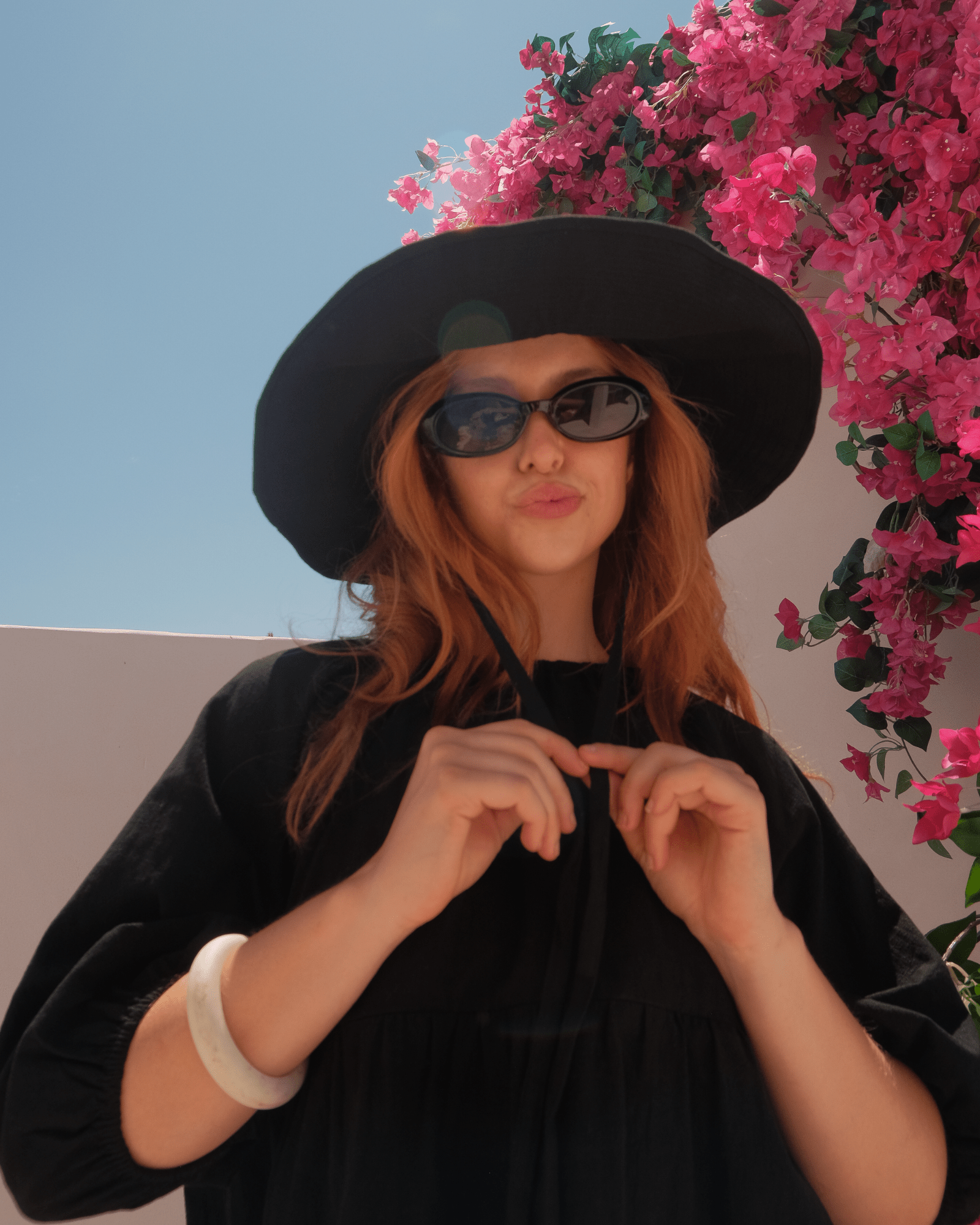 Person wearing a black wide-brimmed hat with tie and sunglasses with pink flowers in the background