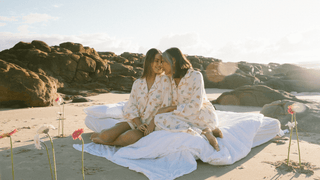 A mother and daughter wearing cosy pyjamas sitting on a bed on a sunny beach in the morning, surrounded by flowers