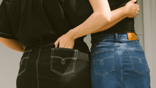 Two women standing next to each other with their hands in each others jean pockets. It's a close up of two pairs of jeans, blue and black.