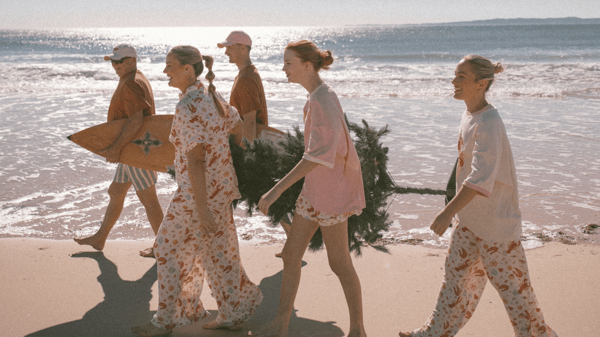 Family walking along the beach wearing christmas pyjamas, holding a christmas tree and surf board.