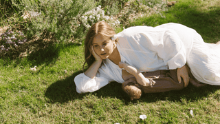 A mother breastfeeding her child laying down in the grass wearing an avalon maxi smock. 