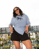 A woman wearing a light blue oversized t-shirt with a black horse graphic and black pleated shorts, posing outdoors on a balcony with a decorative railing.