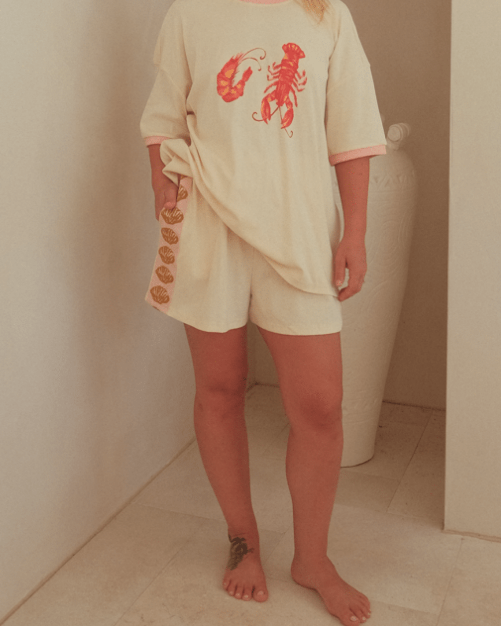 Woman wearing cream coloured summer lounge set with oversized lobster graphic t-shirt and shorts with pink & gold detail down the side-seam. 