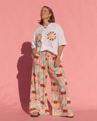 Person wearing a white t-shirt with floral designs and colorful wide-leg pants against a pink background