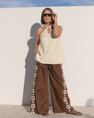 Woman wearing a pale yellow sleeveless top and brown pants with white star patterns against a white wall.